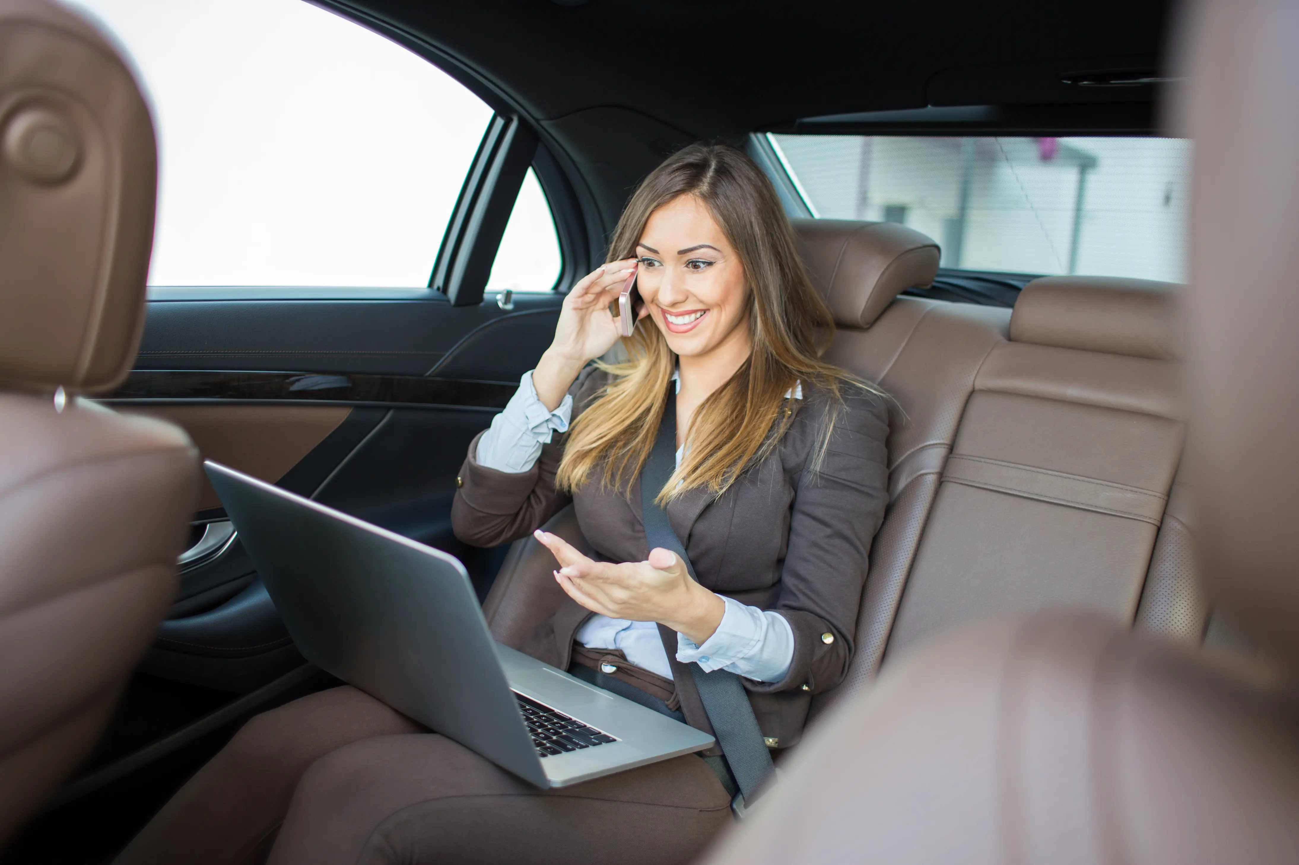 Eine Frau mit Laptop im Schoß und Handy am Ohr auf der Rückbank eines Autos. Der Blick aus dem Fenster ist nicht möglich, da die Scheibe weiß undurchsichtig ist.