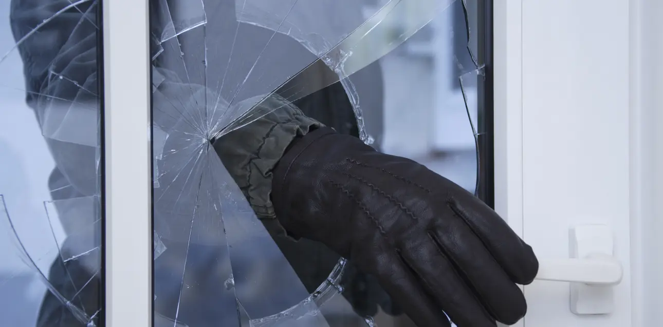 Eine Person greift mit einer Hand im Handschuh durch die zerbrochene Scheibe einer Tür an die Türklinke.